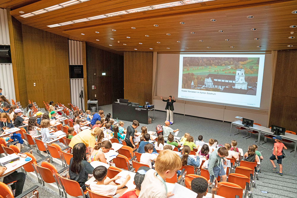 Fast alle orangen Stühle im grossen Vorlesungssaal der Uni Basel sind mit Kindern besetzt. Delpine Conzelmann steht vor einer Leinwand auf der Kloster Mariastein abgebildet ist. Die meisten Kinder sind konzentriert, ein Junge läuft die Stufen hinunter.